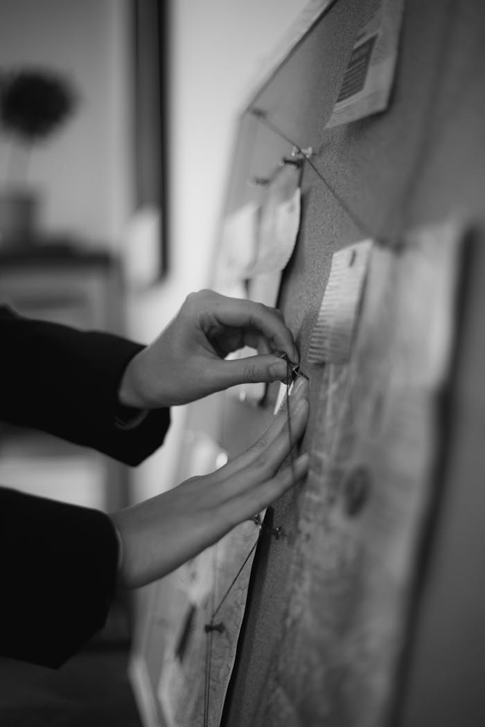 Close-up of hands pinning notes on a cork board in black and white.